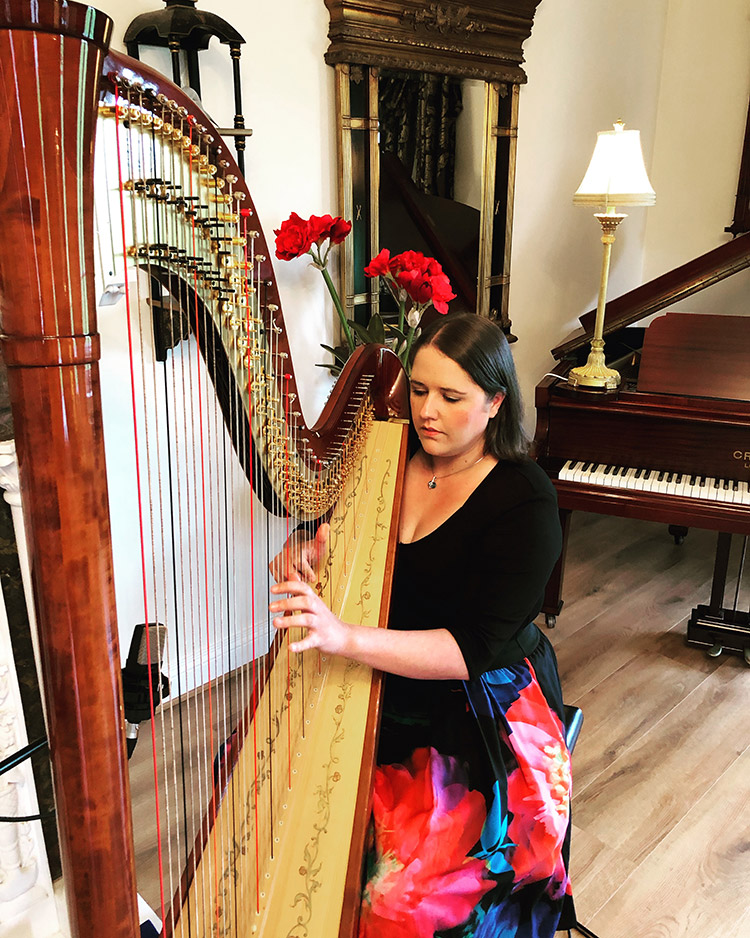 Aisleagh playing harp in a large room with a baby grand piano behind her